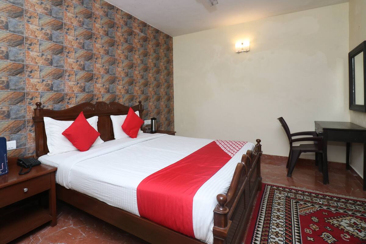 A hotel room with a wooden bed, red and white bedding, a patterned wall, a desk with a chair, and a ceiling fan.