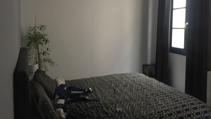 Ett rum med en säng, en krukväxt, ett fönster med gardiner och en vägglampa.