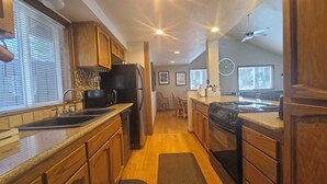 A kitchen with wooden cabinets, a black refrigerator, a stove, and a microwave. There is a window with blinds and a ceiling fan. - 13 Timber-Sunriver, Oregon: Hot Tub, BBQ, WiFi, SHARC Aquatics, Tubing Hill, Mt. Bachelor, Golf, Deschutes River, Tennis, Bike Paths (Sunriver)
