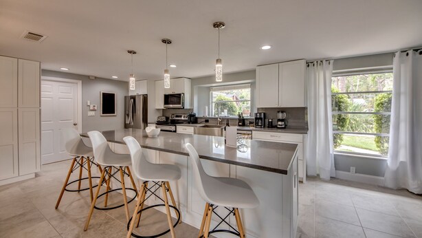 A modern kitchen with white cabinetry, a central island with bar stools, pendant lighting, and a large window with curtains. - Villa Seafan - remodeld, new furniture, heated pool, kayaks, perfect for boaters (North Fort Myers)