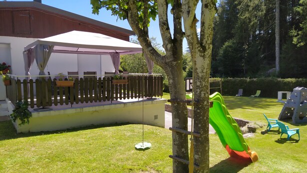 A backyard with a play area including a slide and climbing structure, surrounded by a wooden fence and a white building with a covered patio.