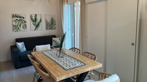 A dining area with a wooden table, wicker chairs, and a decorative runner. There is a sofa with cushions in the background and a door to the right.