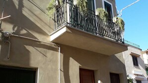 A two-story building with a balcony featuring a wrought iron railing and potted plants. There are two doors, one with a green shutter and another with a wooden shutter.
