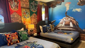 Une chambre d’hôtel à thème, avec une fresque murale représentant Poudlard, deux lits avec une literie assortie au thème, et une table de chevet avec une lampe.