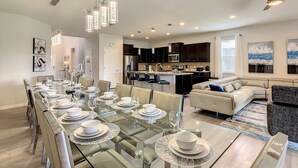 Un espace de repas moderne avec une longue table dressée pour six personnes, une cuisine en arrière-plan et un coin salon avec un canapé et une table basse.