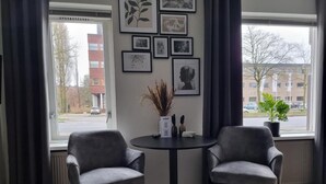 A room with two grey armchairs around a small round table, a window with a view of buildings, and framed pictures on the wall. - Studio Trenz for two - Groningen Center (Groningen)