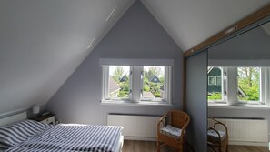 Un dormitorio con techo inclinado, cama con ropa de cama a rayas, una silla de mimbre y vistas a casas vecinas a través de amplios ventanales.