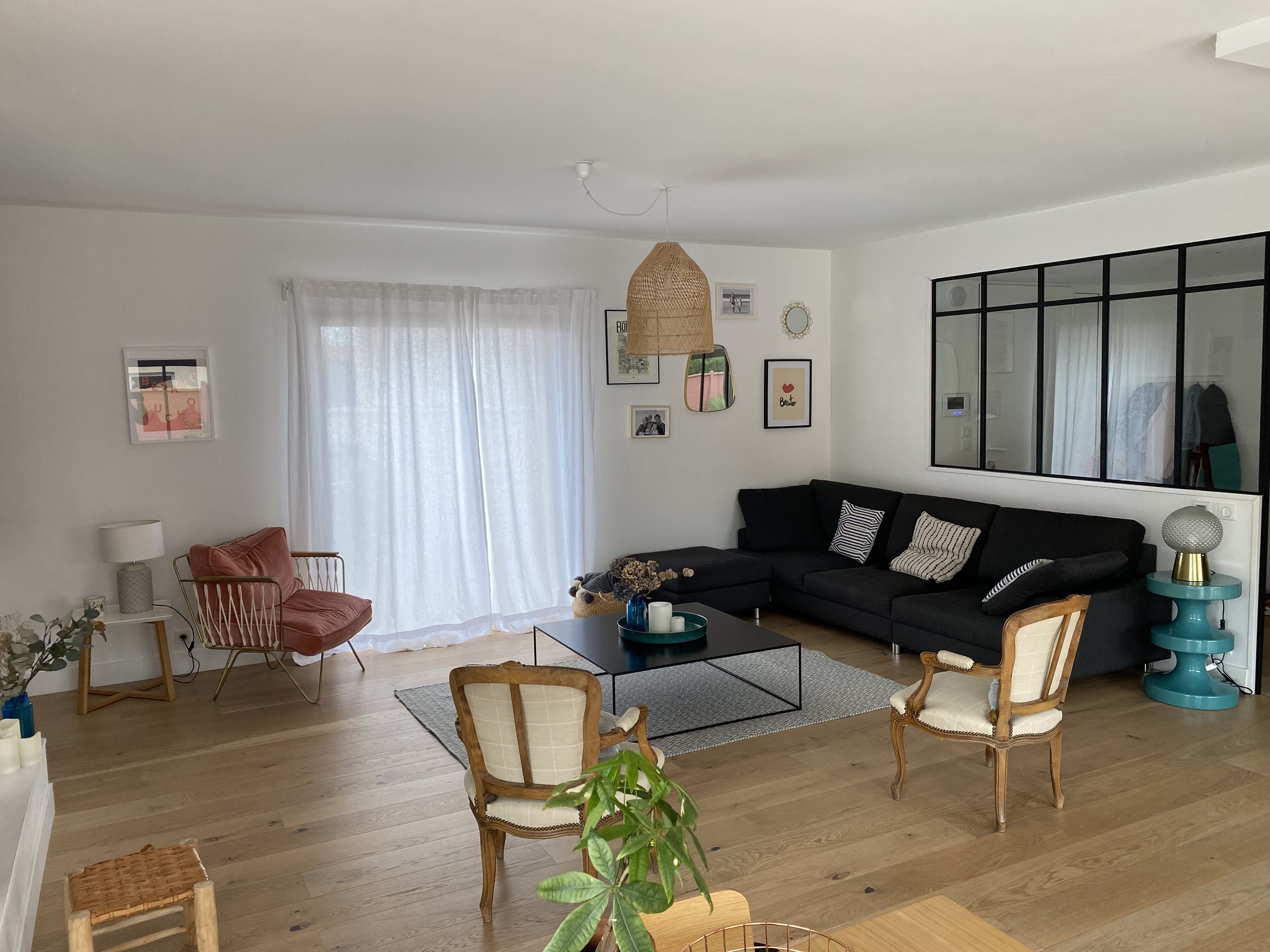 Een moderne woonkamer met een zwarte bank, houten stoelen, een salontafel en een groot raam met transparante gordijnen.