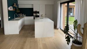 A modern kitchen with white cabinetry, a central island, and a view of a wooden deck through large windows. - Large villa with swimming pool on the outskirts of Bordeaux (BRUGES)