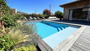A swimming pool with a wooden deck, surrounded by lush greenery and a clear blue sky. - Large villa with swimming pool on the outskirts of Bordeaux (BRUGES)
