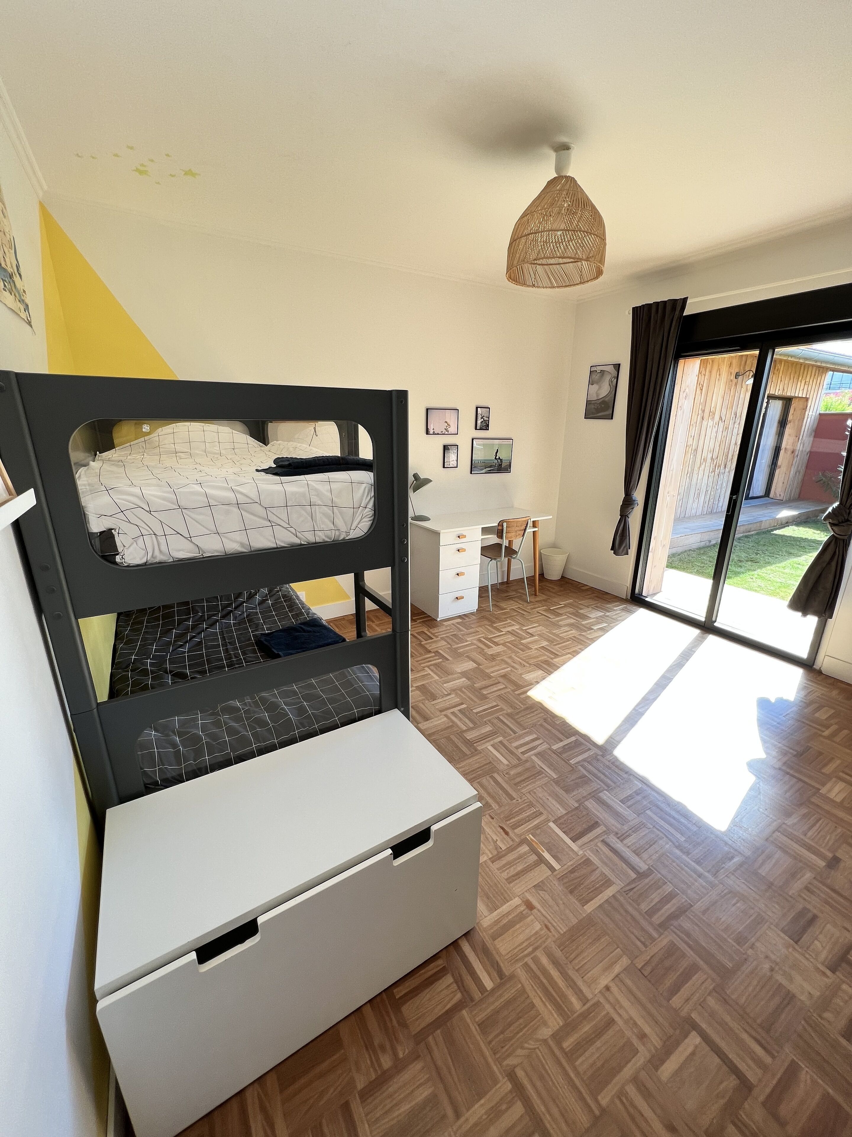 Una stanza con un letto a castello, una scrivania e una sedia. Alle pareti ci sono dei quadri e una finestra con tende.