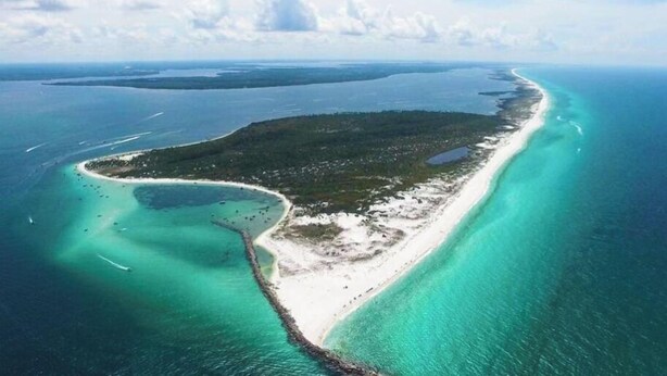 Vista aérea de una isla costera con una playa estrecha, aguas turquesas cristalinas y una zona boscosa densa.