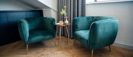 A room with two teal velvet armchairs, a small round table with a vase, and a woven rug on a wooden floor.