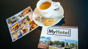A coffee cup with a spoon and a biscuit on a saucer, a brochure titled "MyHotel STEENWUK," and a table with a dark wooden surface. - MyHotelSteenwijk (Steenwijk)