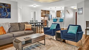Ein modernes Wohnzimmer mit einer Couch, Sesseln und einem Couchtisch. Es gibt einen Essbereich mit Tisch und Stühlen, und im Hintergrund eine Küche.