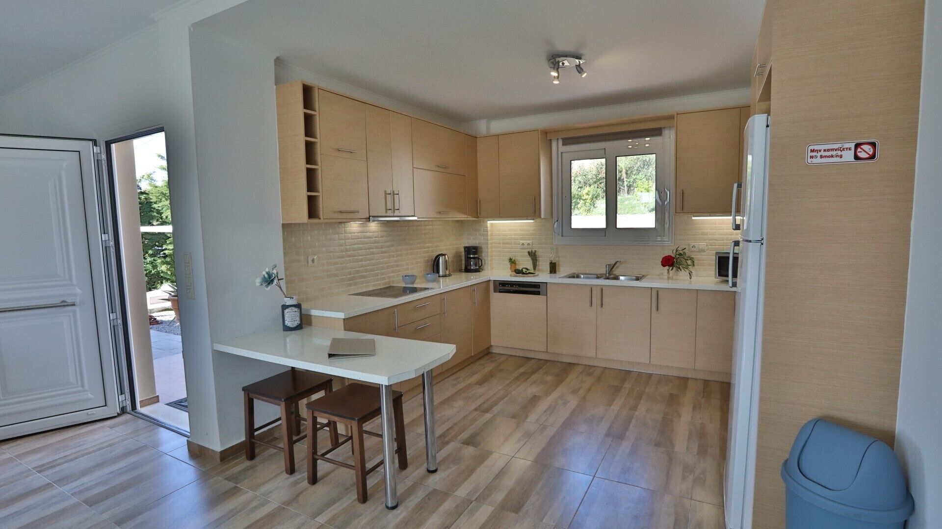 A modern kitchen with light wood cabinets, a white countertop, and a built-in dishwasher. There is a window providing natural light, and a blue trash bin is visible.