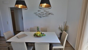 Uma área de refeições com uma mesa branca, quatro cadeiras, duas luminárias pendentes e uma tigela de frutas.