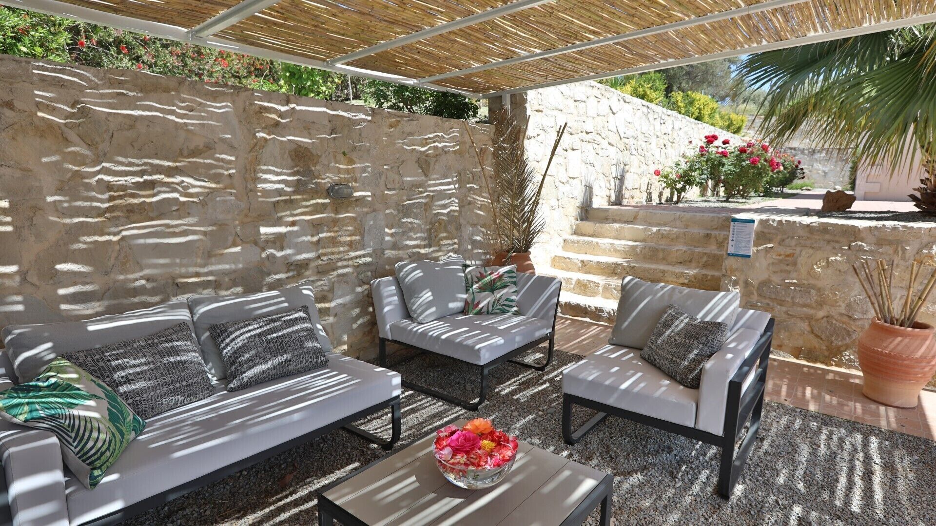 Ein Außenbereich zum Sitzen mit Steinmauern, einer Pergola und einem kleinen Tisch mit Blumen.