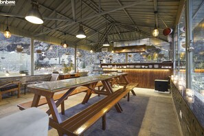 Un restaurant avec des tables et des bancs en bois, des murs en pierre et de grandes fenêtres donnant sur un paysage de montagnes enneigées.