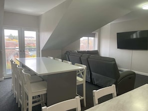 Ein modernes Zimmer mit einem langen Tisch und Stühlen, einem grauen Sofa, einem Flachbildfernseher an der Wand und großen Fenstern mit Blick nach außen.