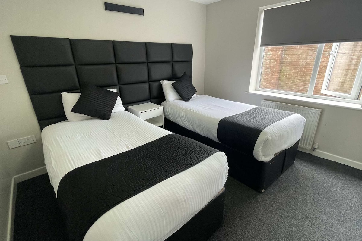 A hotel room with two beds, a black headboard, white bedding, a window with blinds, and a radiator.