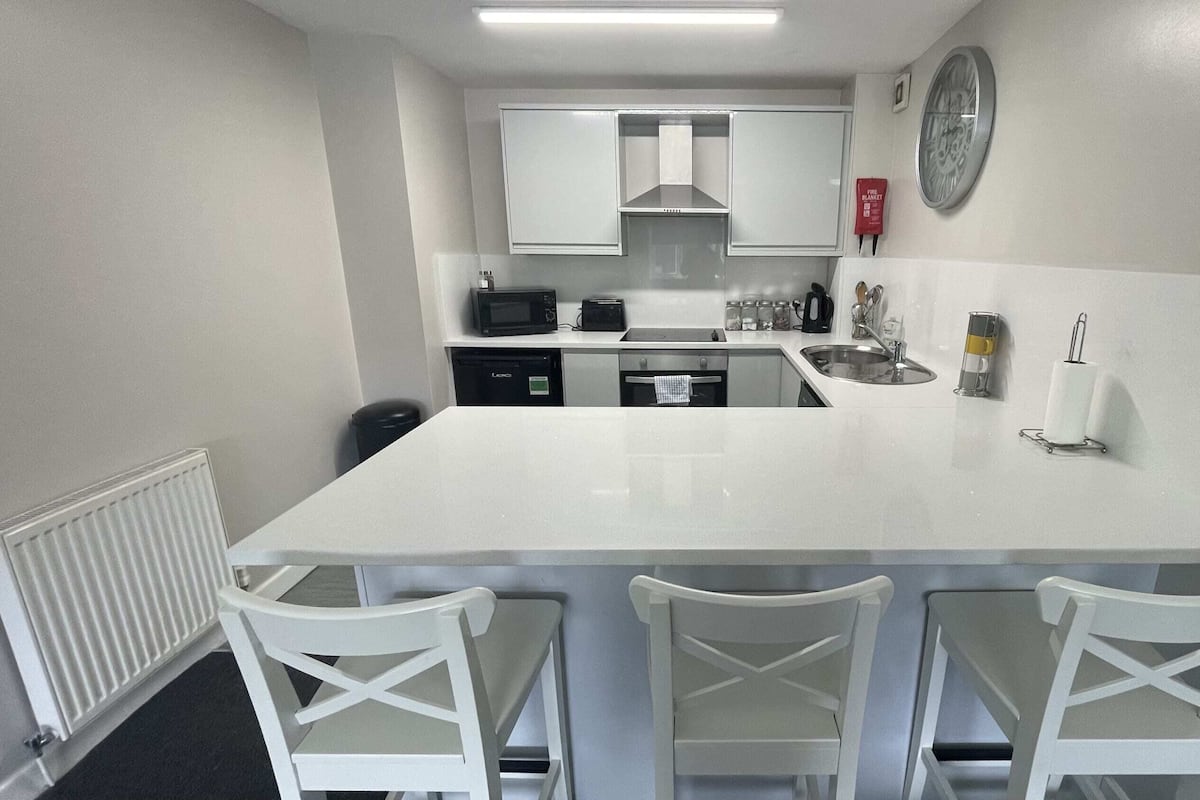 A compact kitchen with a central island, white chairs, a microwave, a toaster, and a clock on the wall.