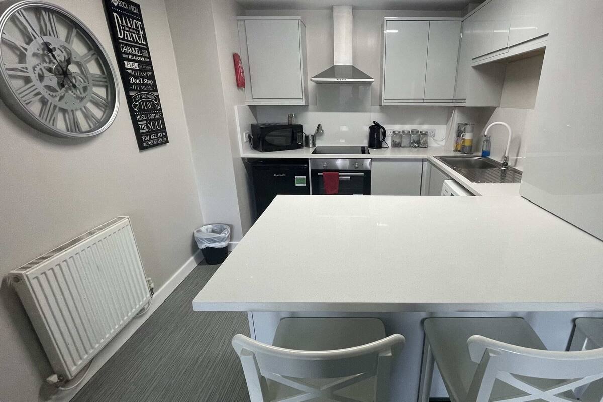 A compact kitchen with white cabinets, a microwave, a stove, and a sink. There is a clock on the wall and a trash bin near the door.