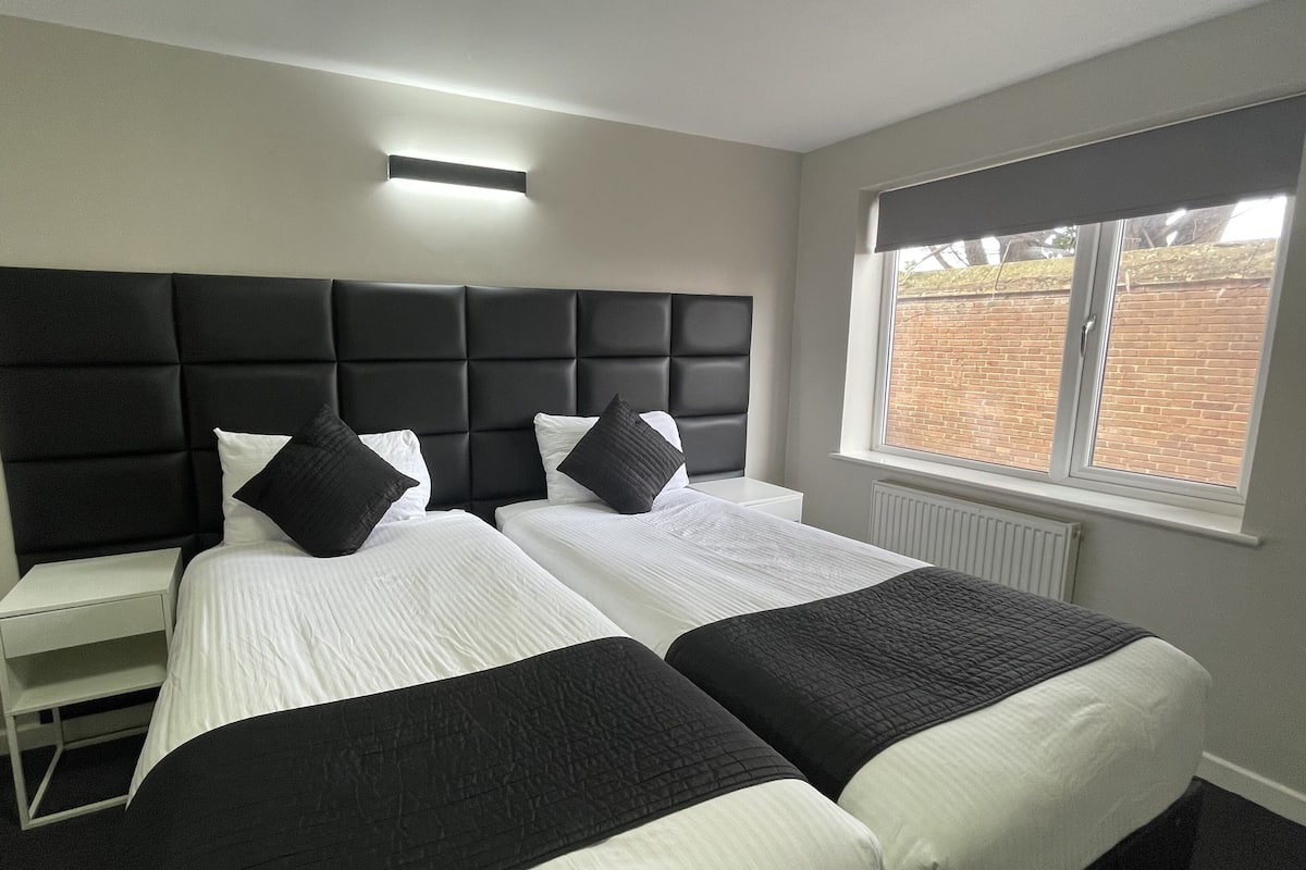 A hotel room with two beds, a large padded headboard, a bedside table, a window with a view of a brick wall, and a ceiling light.
