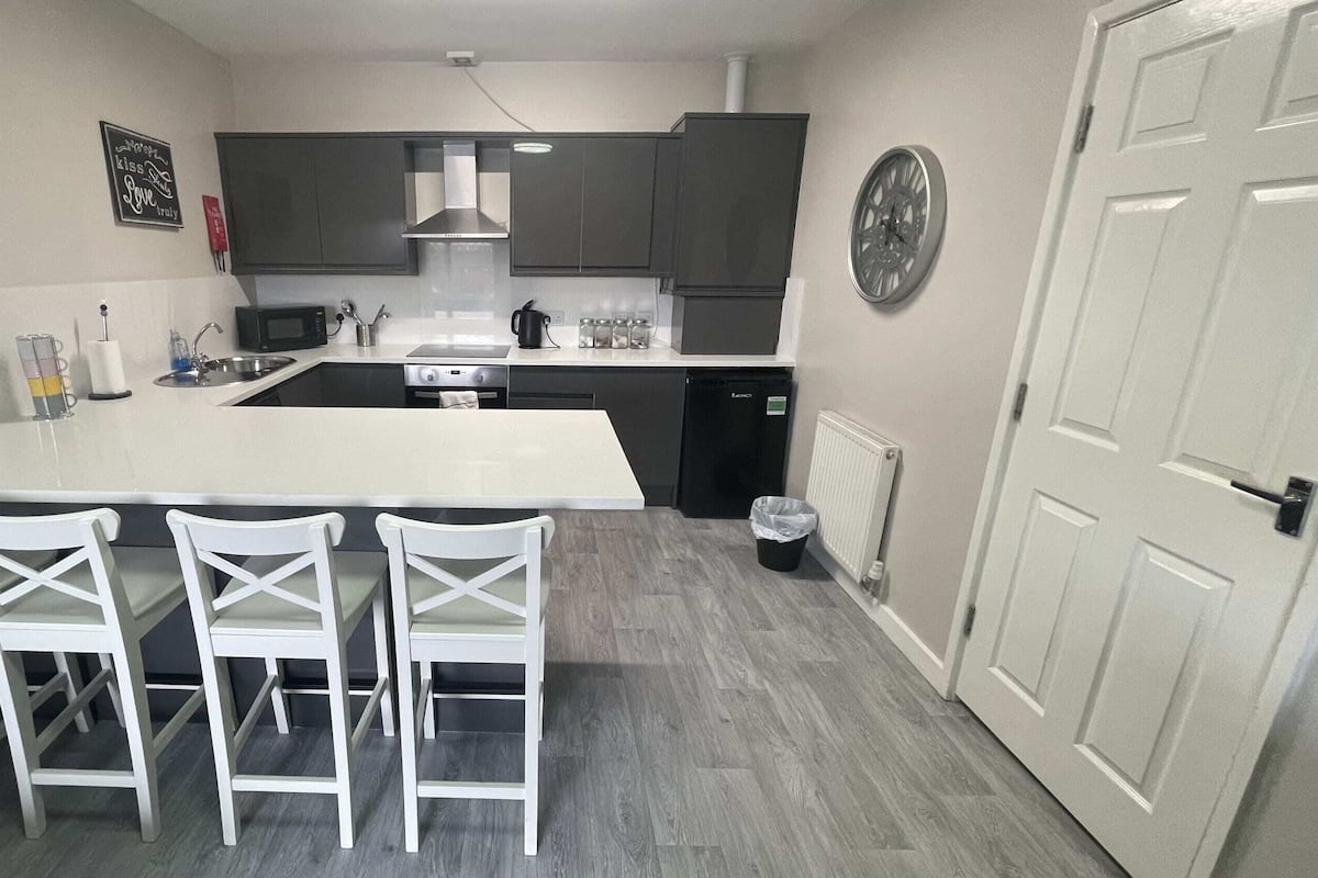 A compact kitchen with a central island, four white chairs, dark cabinets, a microwave, a kettle, and a clock on the wall.