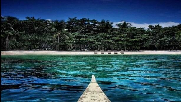 Un muelle de madera se extiende hacia aguas turquesas cristalinas, que conducen a una playa de arena blanca y un denso follaje tropical al fondo.