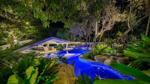 Vista nocturna de un área de piscina con una zona de estar techada y abundante vegetación.