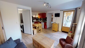 En stue med sofa, sofabord i træ og en rød lænestol. Der er et køkkenområde med røde skabe og et spisebord. Et fjernsyn er monteret på væggen, og en skydedør fører til et udendørs område.