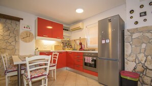A kitchen with red cabinets, a stainless steel refrigerator, a stone wall, and a dining area with white chairs. - Apartment Maja 2 (Premantura)
