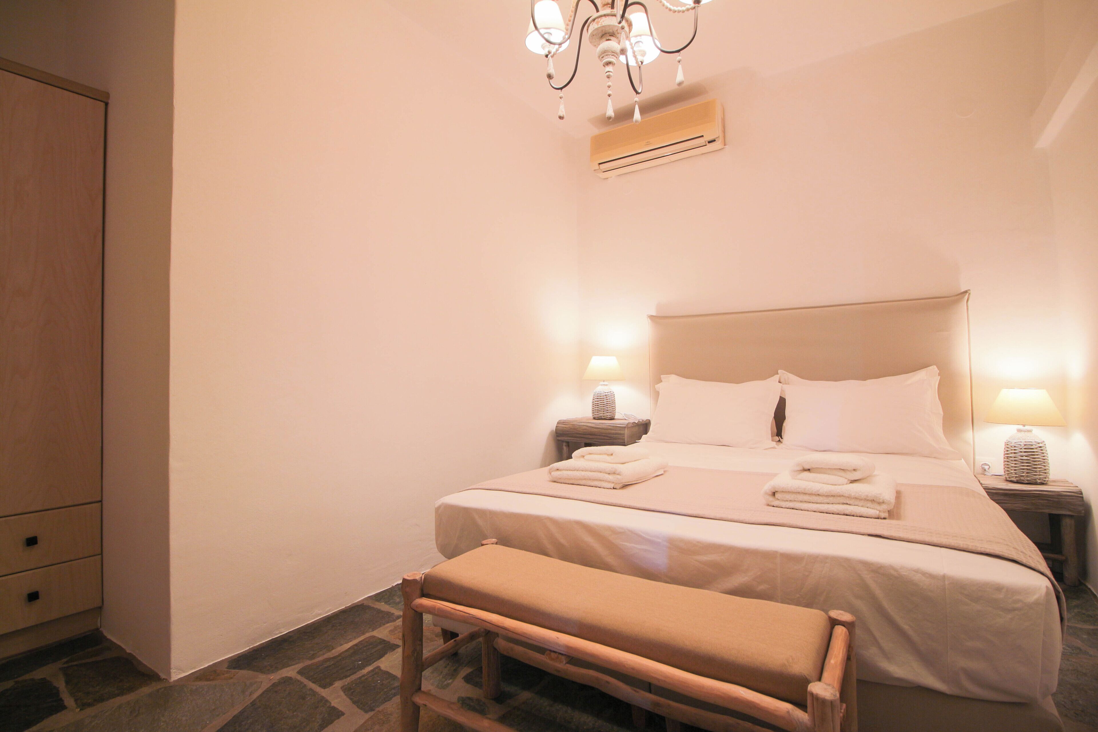 Cama bem arrumada com lençóis brancos, um banco, um candelabro e um ar condicionado fixado na parede.