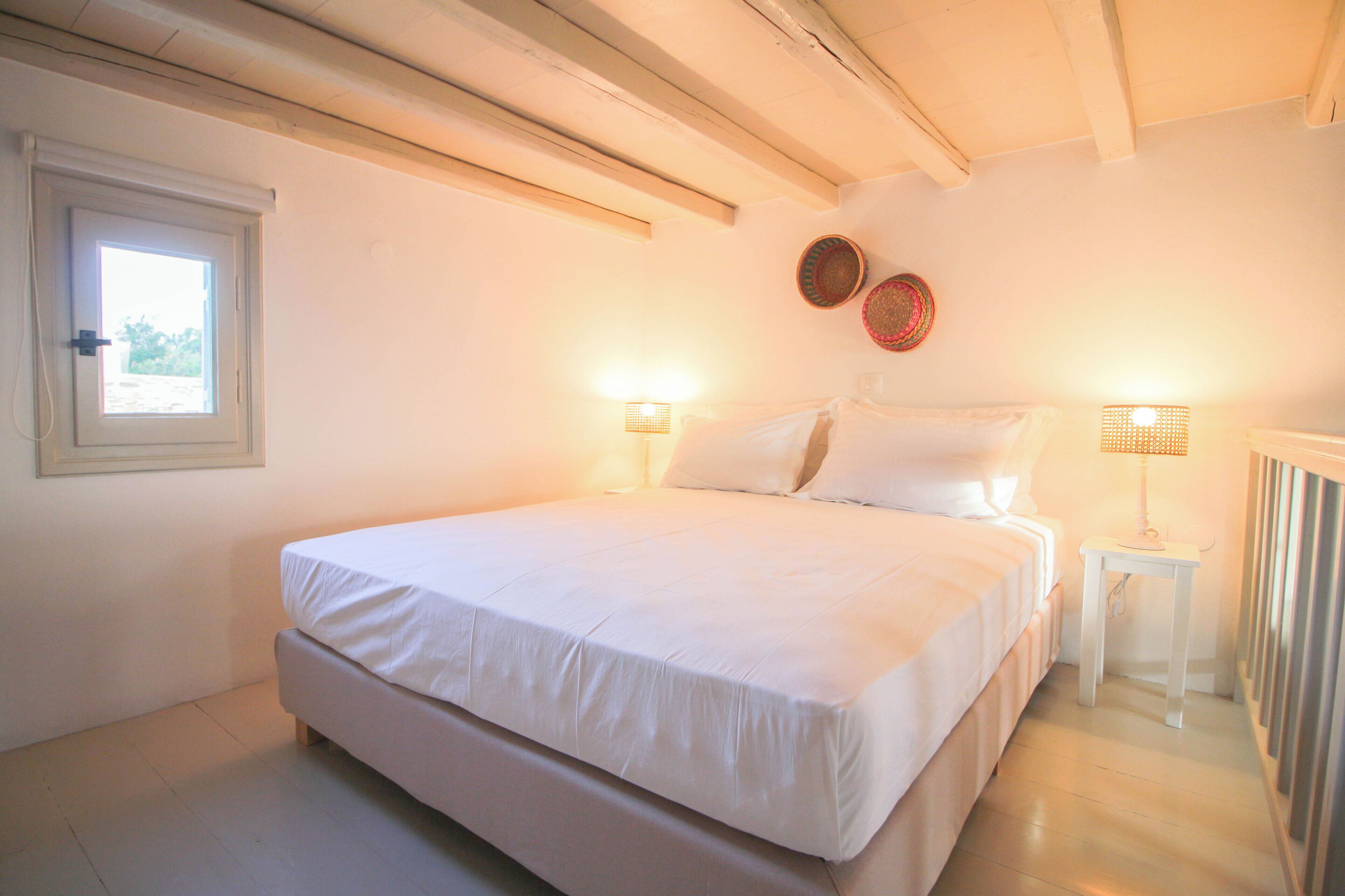 Quarto com uma cama grande, duas luminárias de cabeceira, uma mesinha e uma janela.