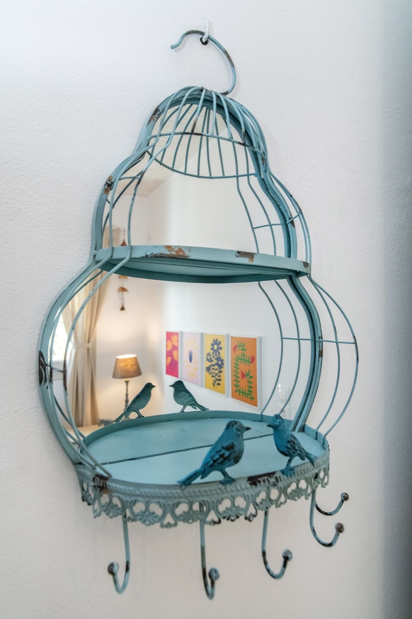 Una estantería azul en forma de jaula de pájaros con ganchos, dos pájaros y un cuadro en la pared de fondo.