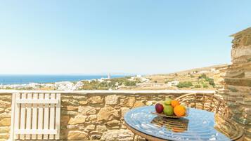 Un balcón con pared de piedra, una mesa de mimbre con fruta y vista al mar y edificios.