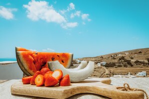 Una tabla de madera con sandía y fresas en una playa, con casas al fondo.