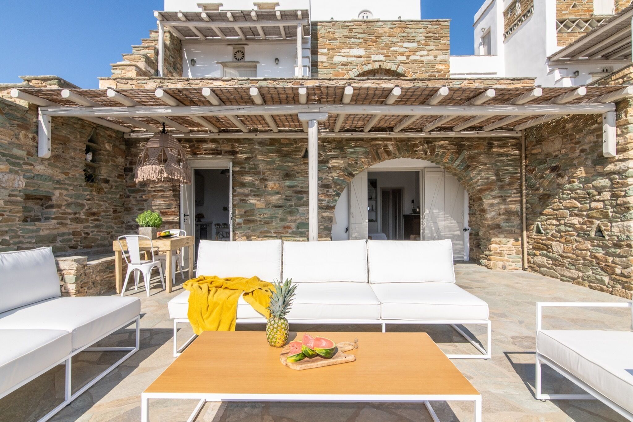An outdoor seating area with white sofas and a wooden table, stone walls, and a pergola.