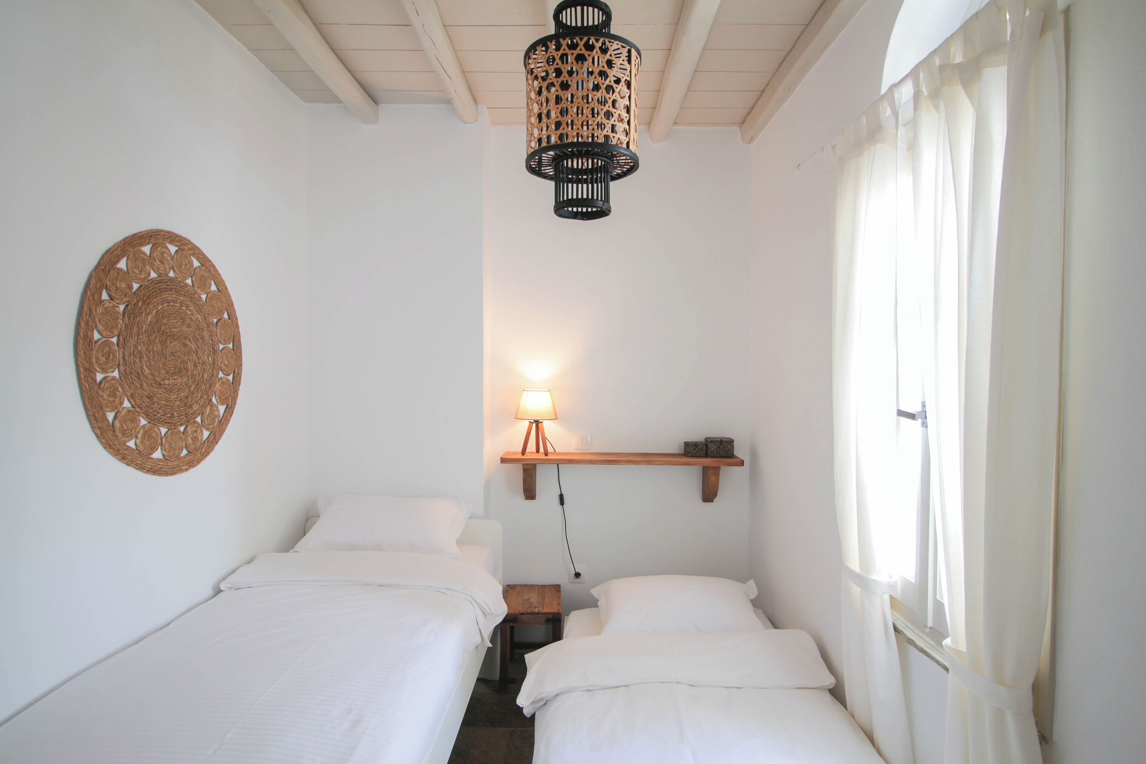 A small, well-lit bedroom with two beds, a hanging light fixture, a wall hanging, and a window with curtains.