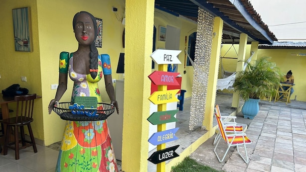 A colorful statue of a woman holding a basket, a yellow wall, and a signpost with multiple directional signs. - Mananaue Villa (Ipojuca)