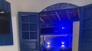 A bedroom with a bed, a nightstand, a mirror, and a view of an outdoor area with blue lighting. - Mananaue Villa (Ipojuca)