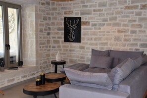 Un salon chaleureux avec un mur en pierre, un canapé gris, deux petites tables rondes en bois et une porte vitrée donnant sur l’extérieur.