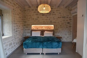 Une chambre avec un mur en pierre, un lit recouvert d’une couvre-lit turquoise, un luminaire suspendu et une fenêtre sur le côté gauche.