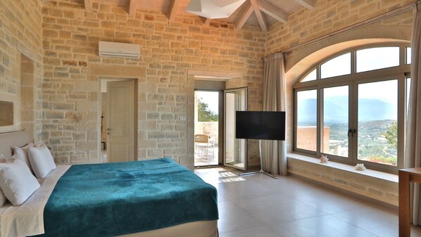 Une chambre avec un mur en pierre, un lit recouvert d’une housse turquoise, une télévision et une grande fenêtre donnant sur un beau paysage.