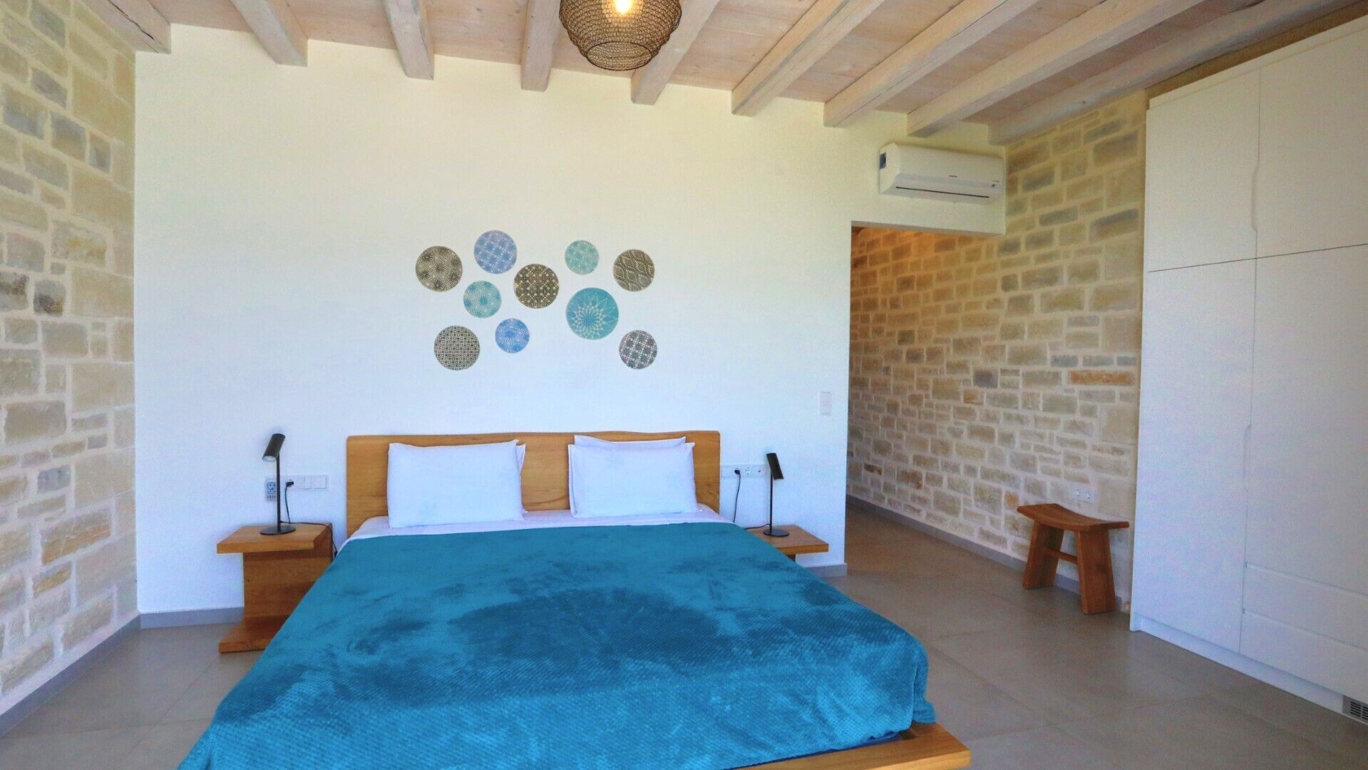 A bedroom with a bed, two bedside tables, a wall-mounted air conditioner, and a stone wall.