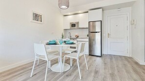 A modern kitchen with white cabinetry, a stainless steel refrigerator, and a dining area with white chairs and a table set for two.