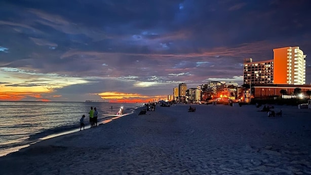 Une plage avec une jetée qui s’avance dans l’eau, un hôtel à plusieurs étages doté de balcons, et un coucher de soleil en arrière-plan.