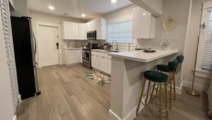 Una cocina moderna con armarios blancos, un refrigerador de acero inoxidable, un microondas y un lavamanos. Hay una pequeña isla con taburetes de bar y un espejo decorativo.