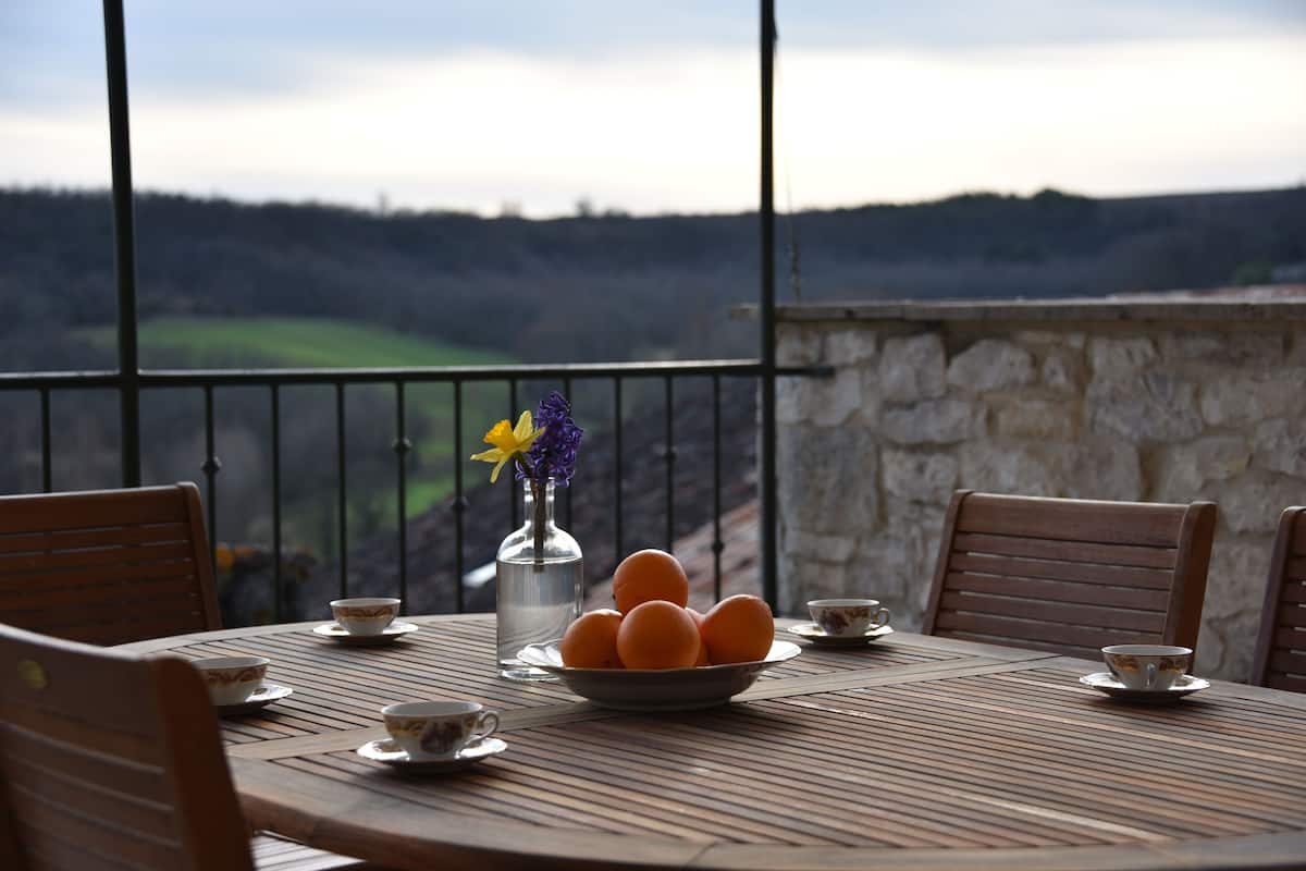 Uma mesa de madeira redonda com uma tigela de laranjas, um vaso de flores e xícaras de chá.